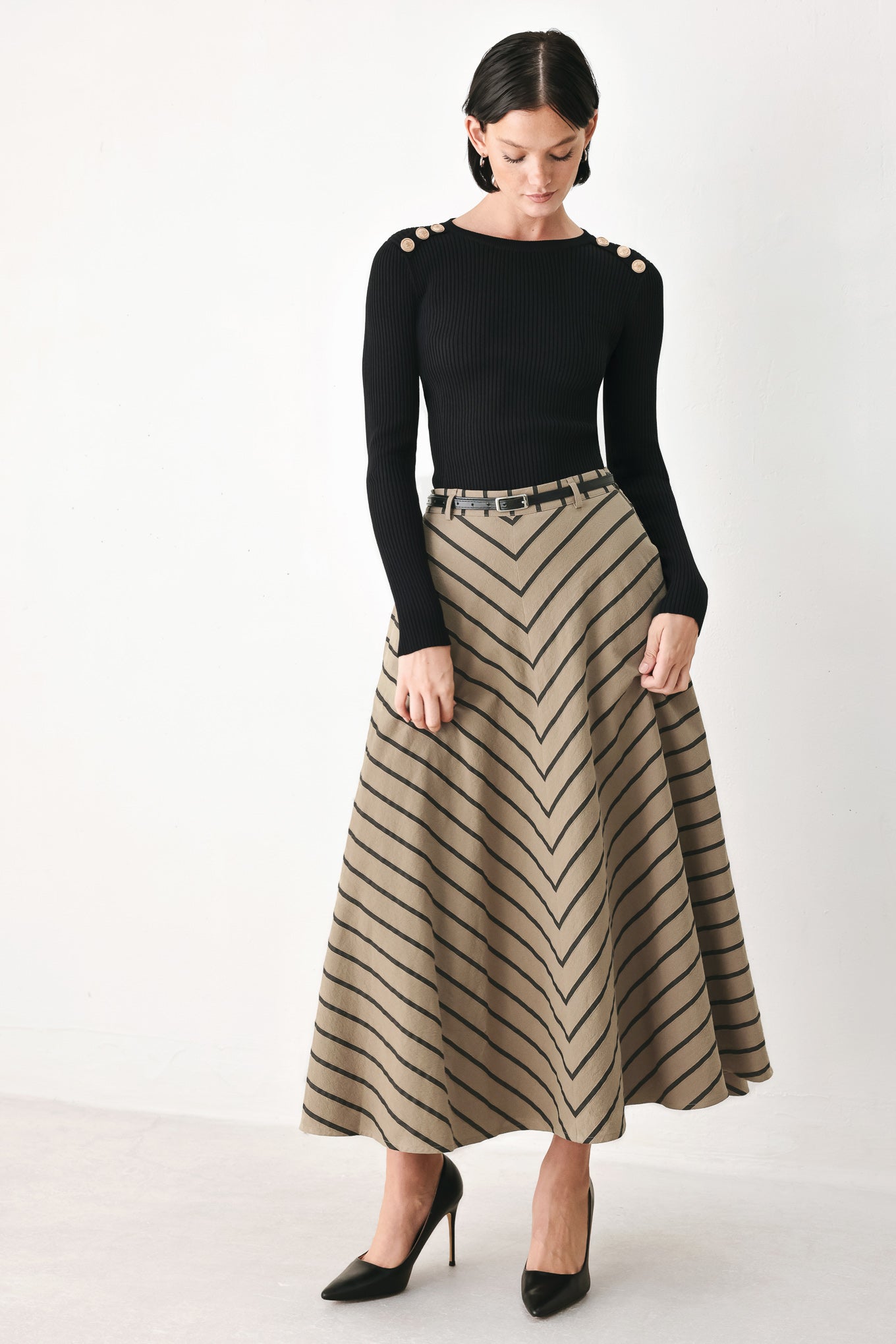 A woman modeling a black long-sleeve top with decorative gold buttons on the shoulders and a high-waisted, flared beige skirt with a black chevron pattern and a black belt. She is wearing black pointed-toe heels against a plain white studio background.