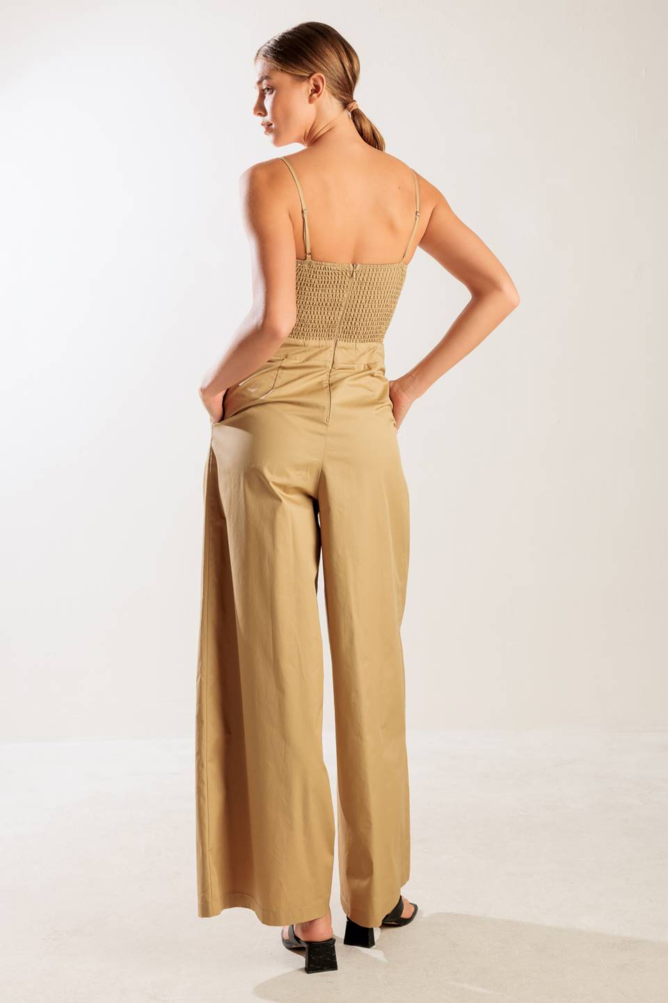 A solid woven jumpsuit featuring straight neckline, smocked bodice, straps, side pockets, wide leg, and back zipper closure