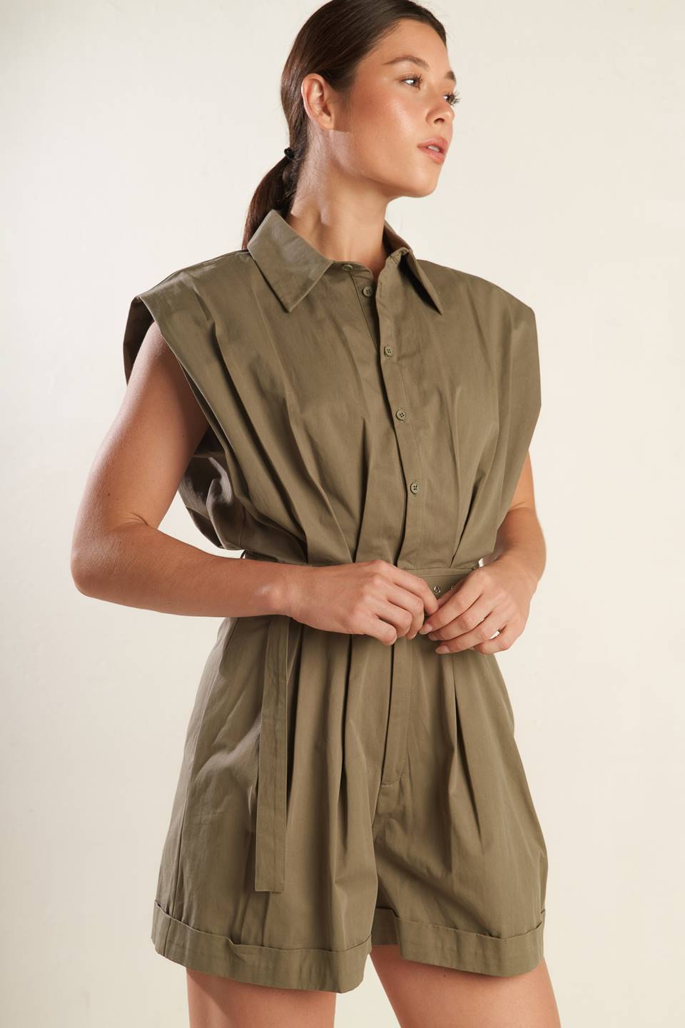 A solid olive woven romper featuring collar, oversize shoulder, front button down and zipper closure, self belt, side pockets and cuffed hemline.