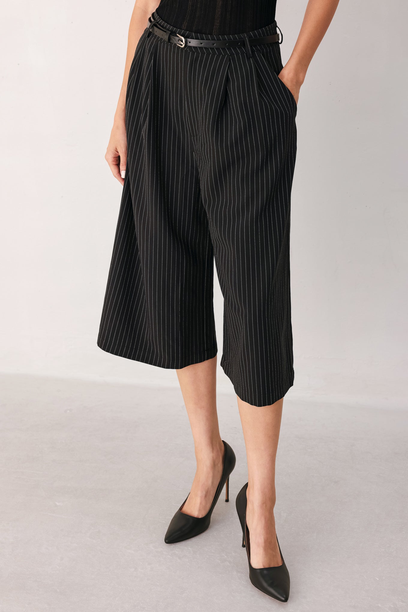 A woman wearing black cropped trousers with thin white vertical pinstripes, featuring a matching black belt with a silver buckle | FREEMARKET