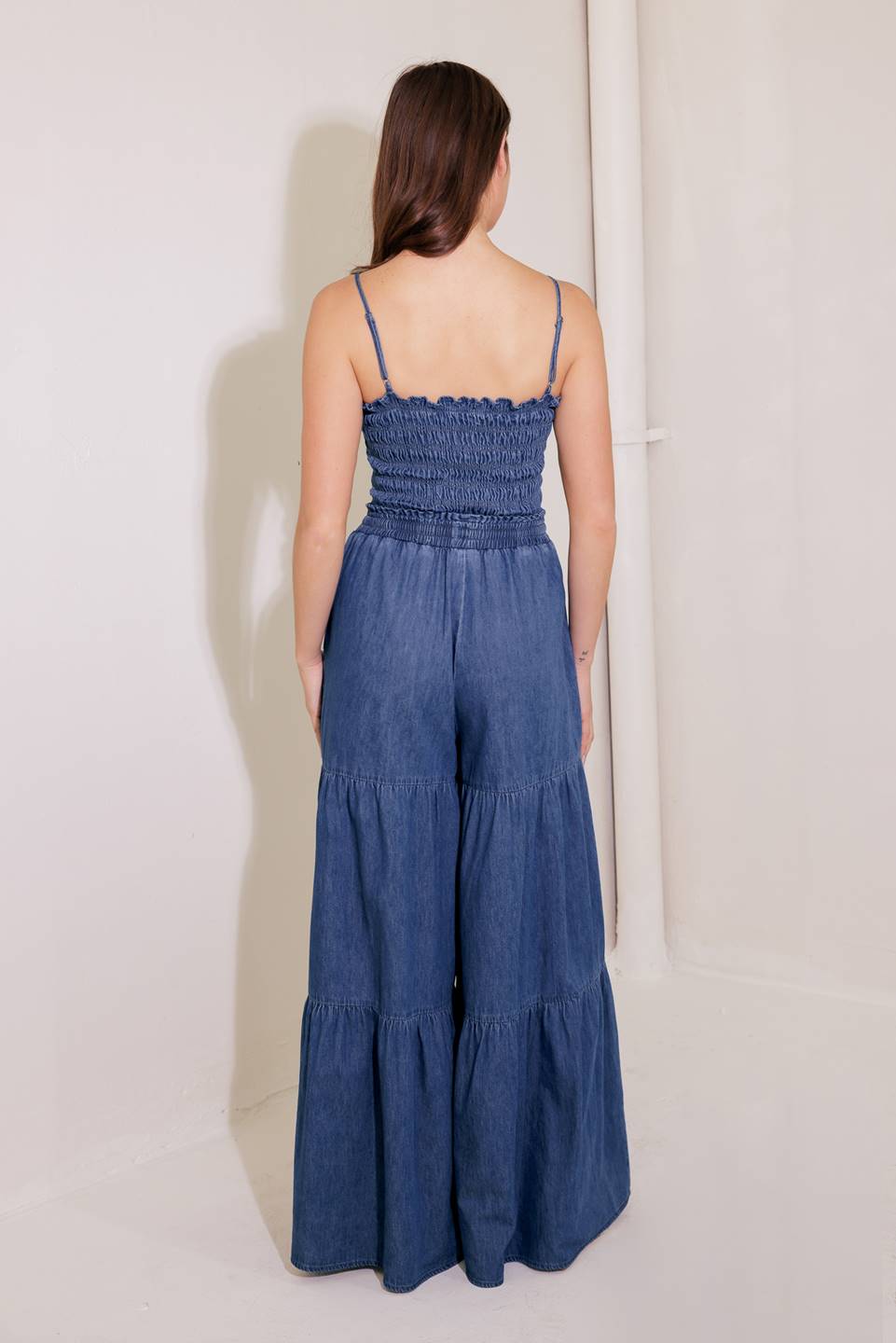 A washed denim jumpsuit featuring strapless, smocked bodice and waistband with front tie and tiered bottom