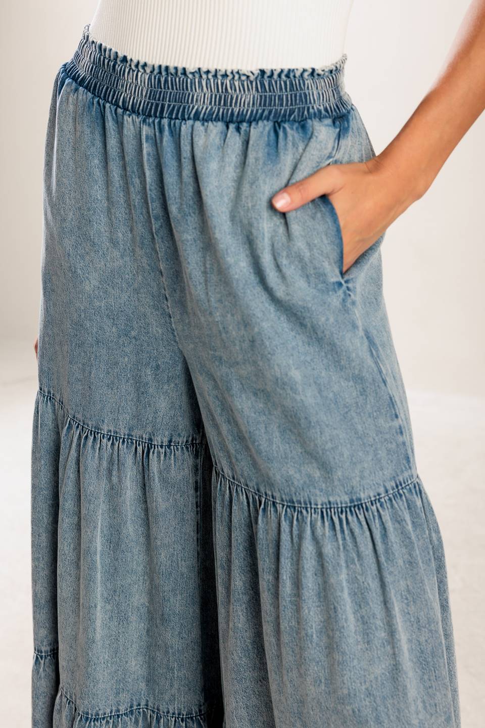 A wide-leg washed denim pant featuring a smocked waistband and tiered bottom.