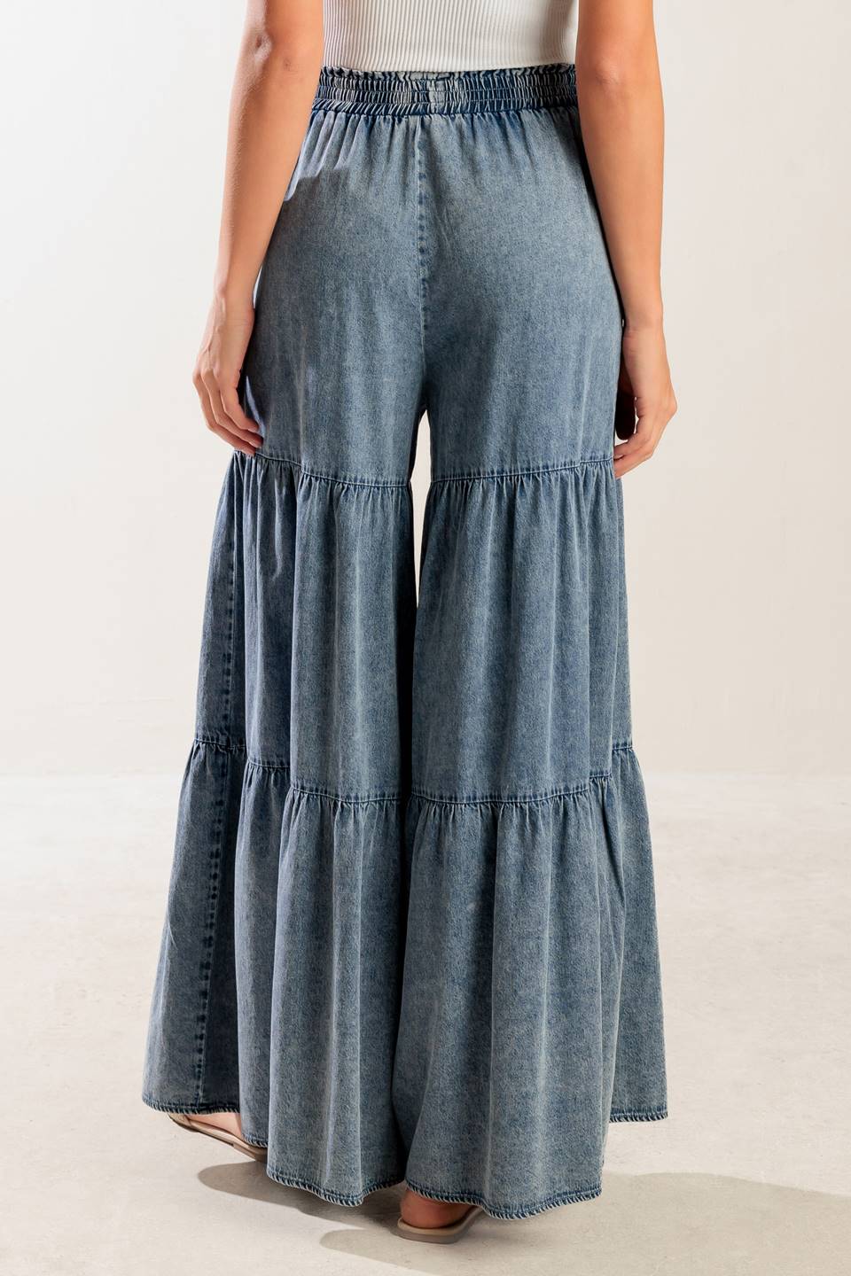 A wide-leg washed denim pant featuring a smocked waistband and tiered bottom.