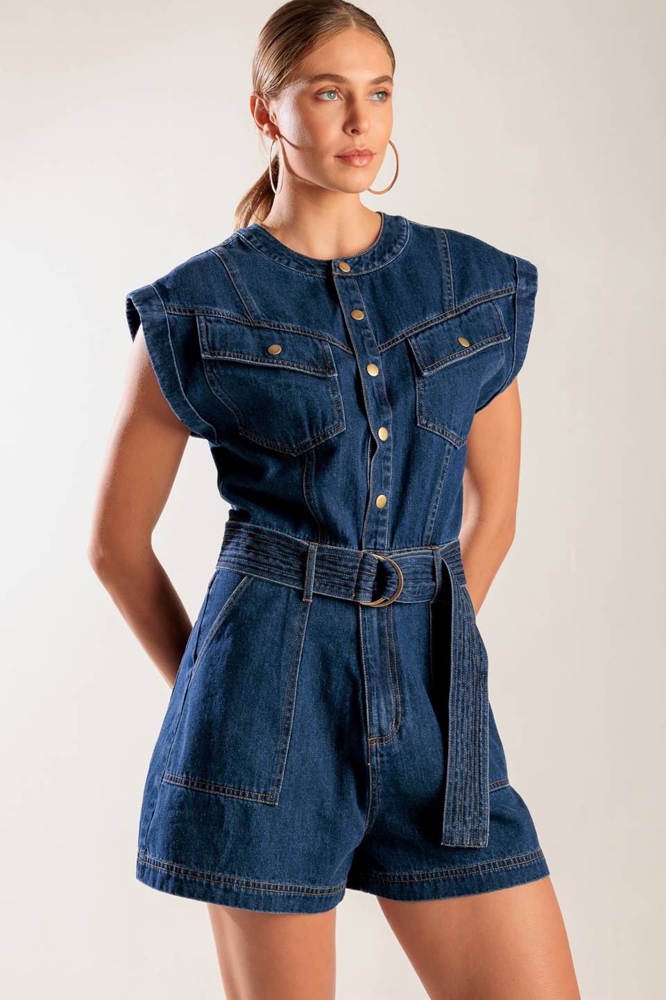 A denim romper featuring a round neckline, button down, cuffed sleeve, self belt, and pockets.