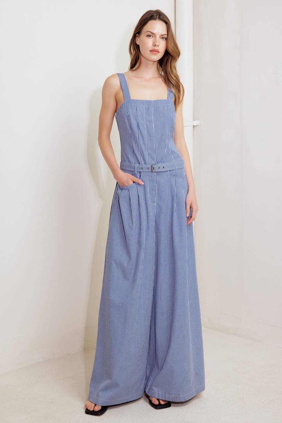 A striped woven jumpsuit featuring straight neckline, straps, front closure, self belt, side pockets and wide leg.