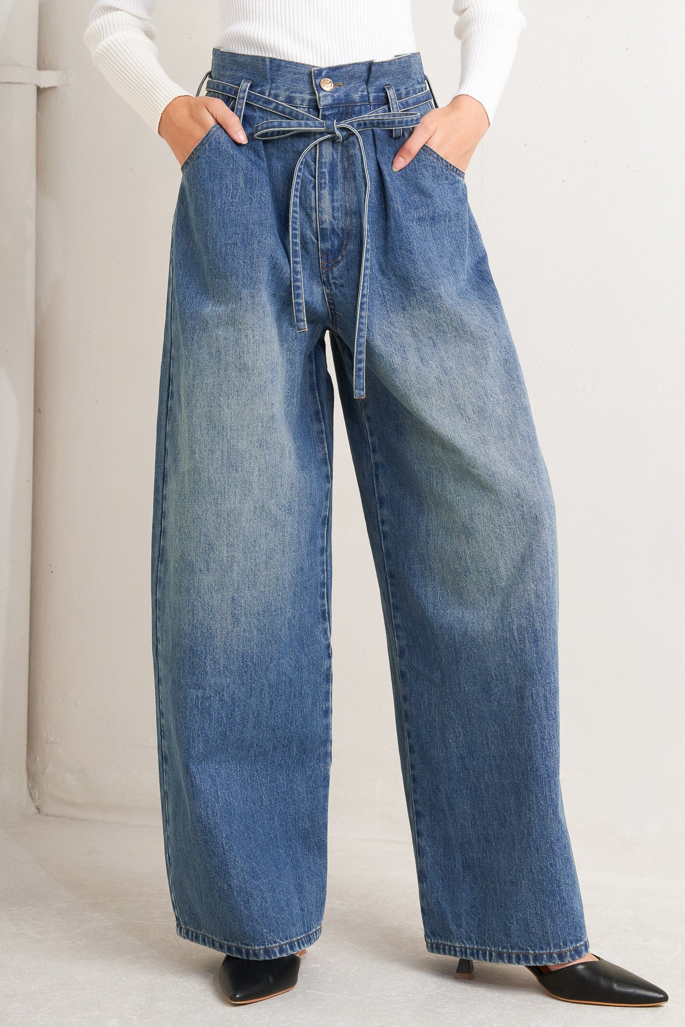 A washed denim pant featuring paper bag waist, front closure, skinny self sash tie, side/back pockets and balloon leg