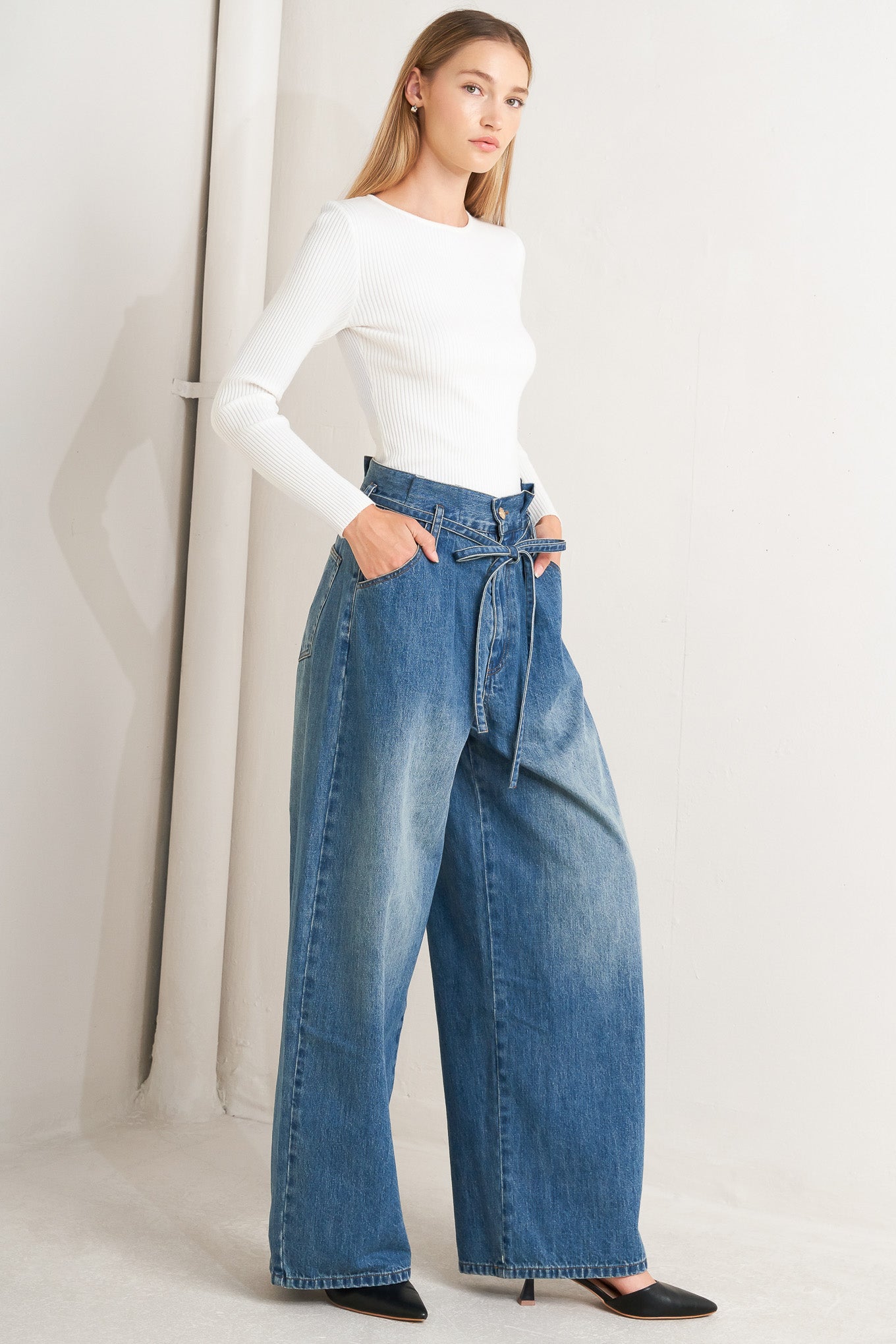 A washed denim pant featuring paper bag waist, front closure, skinny self sash tie, side/back pockets and balloon leg