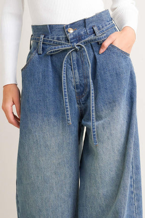 A washed denim pant featuring paper bag waist, front closure, skinny self sash tie, side/back pockets and balloon leg