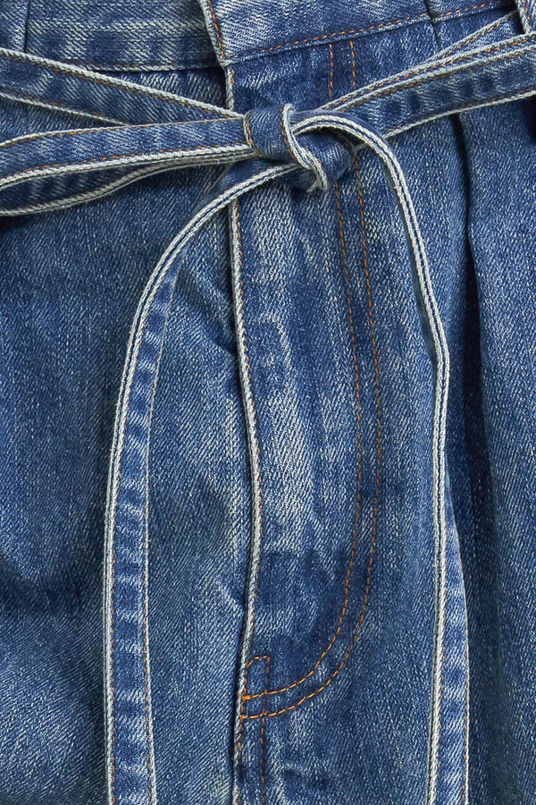 A washed denim pant featuring paper bag waist, front closure, skinny self sash tie, side/back pockets and balloon leg