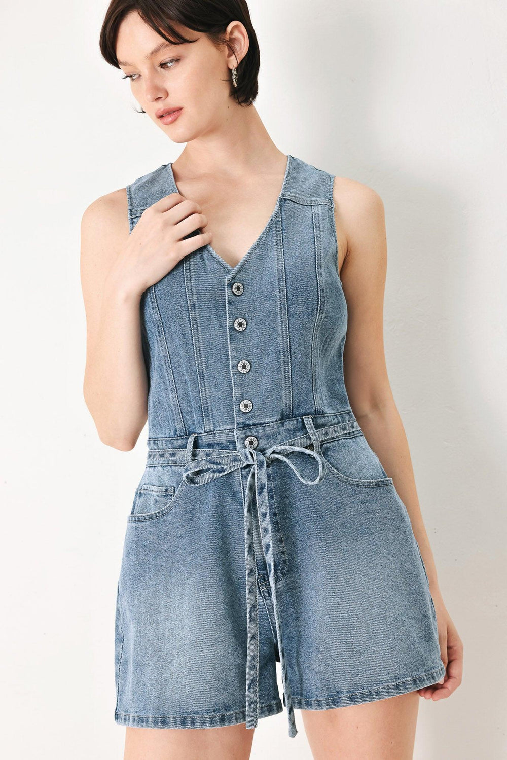 A washed denim romper featuring V neckline, sleeveless, front button/zipper closure, pockets and skinny waist tie