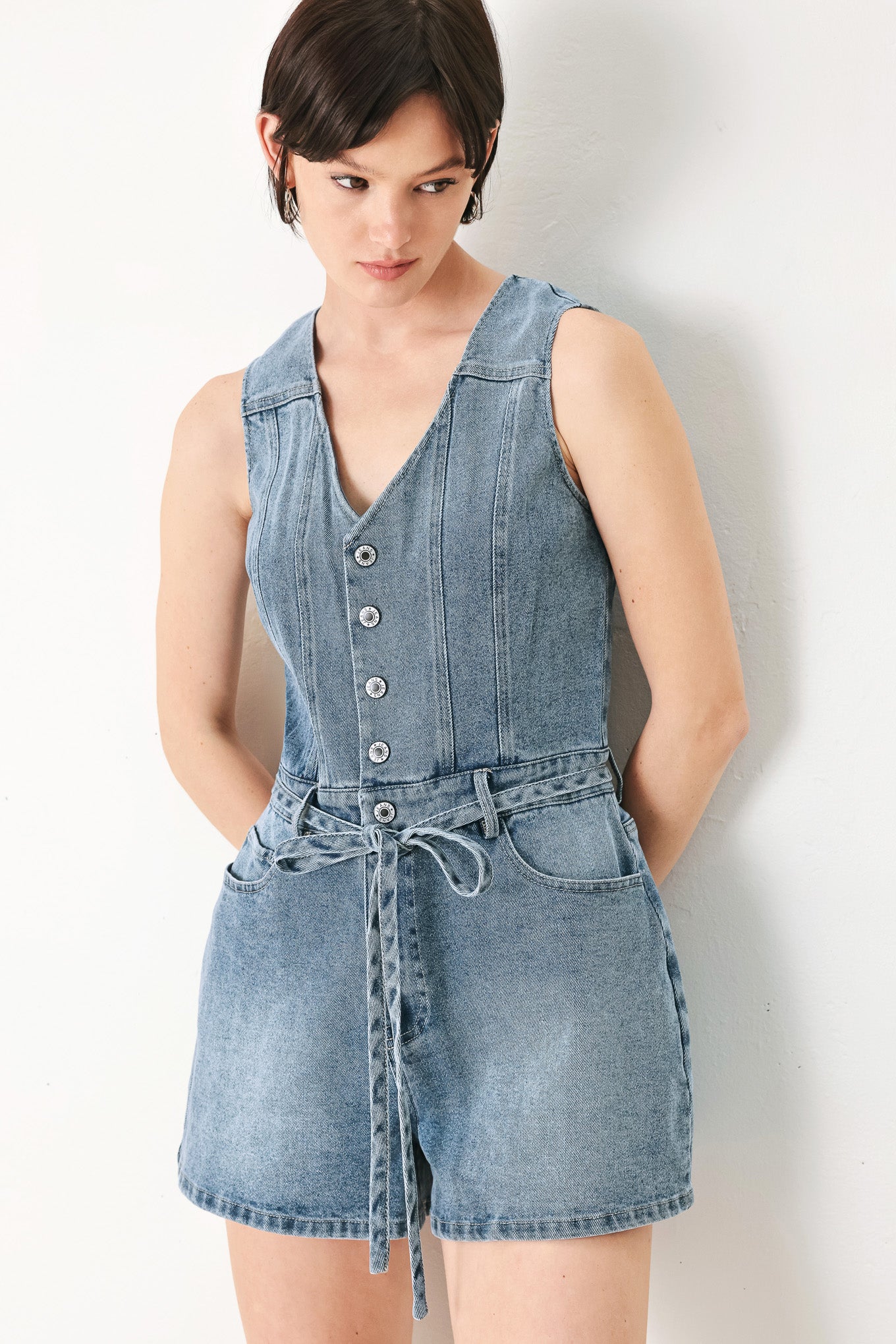 A washed denim romper featuring V neckline, sleeveless, front button/zipper closure, pockets and skinny waist tie