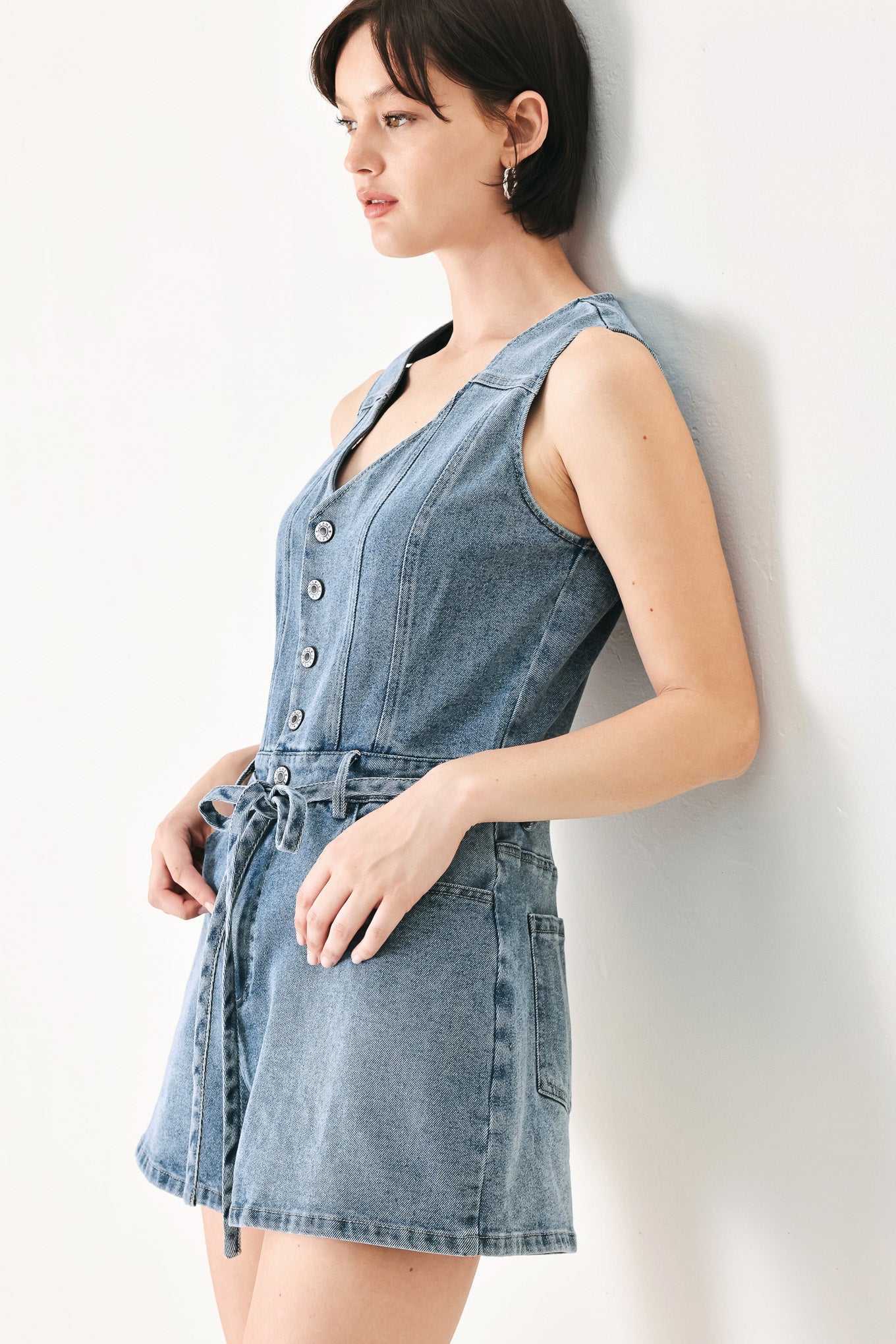 A washed denim romper featuring V neckline, sleeveless, front button/zipper closure, pockets and skinny waist tie