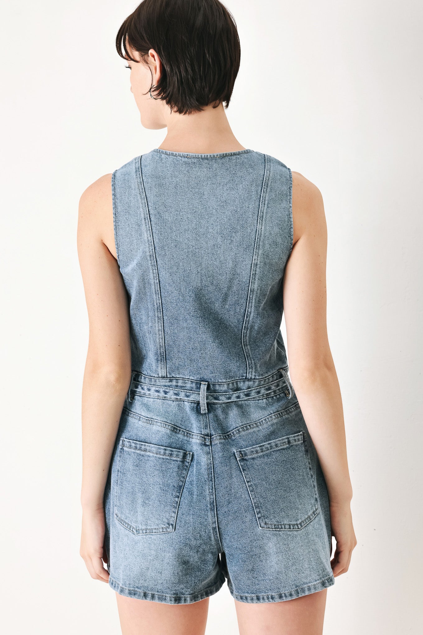 A washed denim romper featuring V neckline, sleeveless, front button/zipper closure, pockets and skinny waist tie