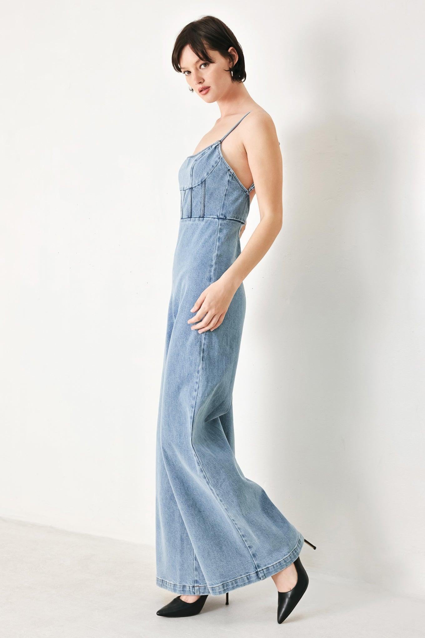 A washed denim jumpsuit featuring U neckline, straps, waist yoke, wide leg, bare back with zipper closure
