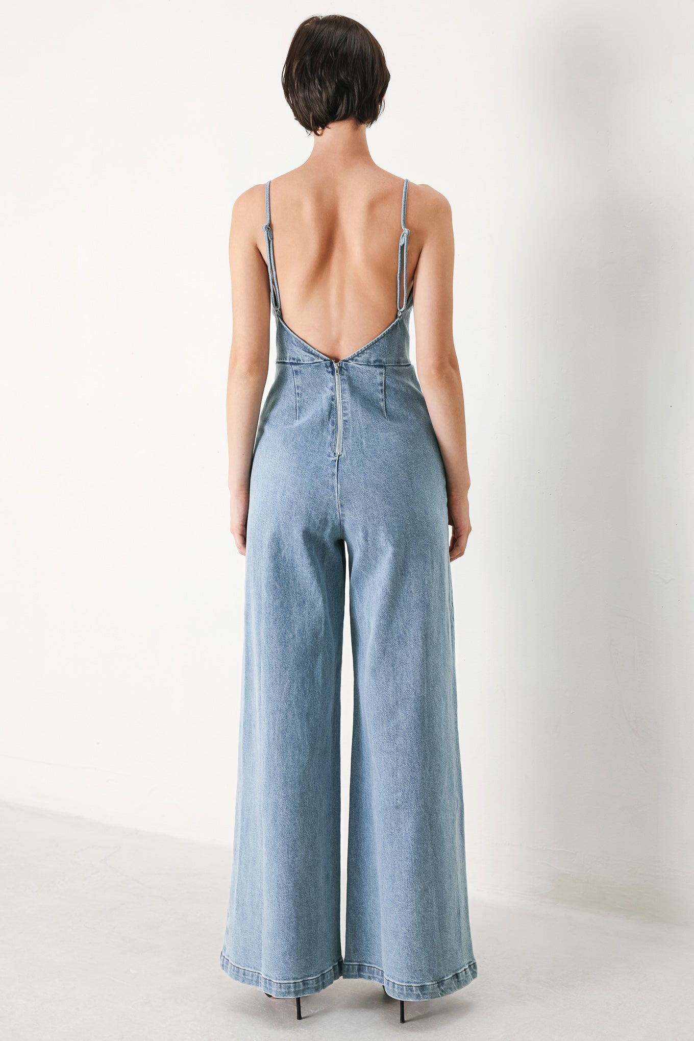 A washed denim jumpsuit featuring U neckline, straps, waist yoke, wide leg, bare back with zipper closure