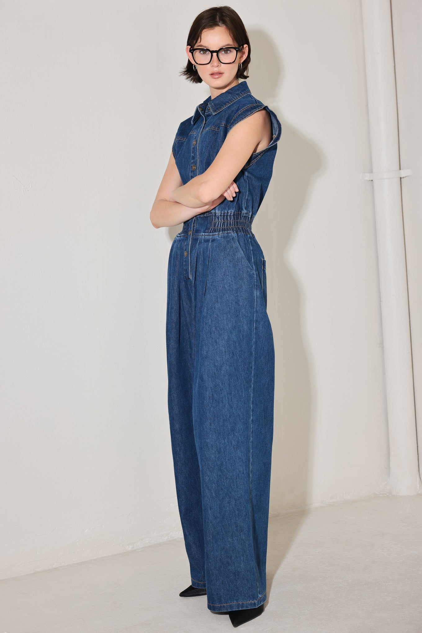 
A washed denim jumpsuit featuring collar, short sleeve, front button down, smocked waistband, side pockets, front pleats and wide leg

Details:

Self : 80% Cotton 20% Polyester

Size &amp; Fit

- Model is 5`8" And Wearing Size Small
- Measurements Taken From Size Small
- Approx. Length: 60"
