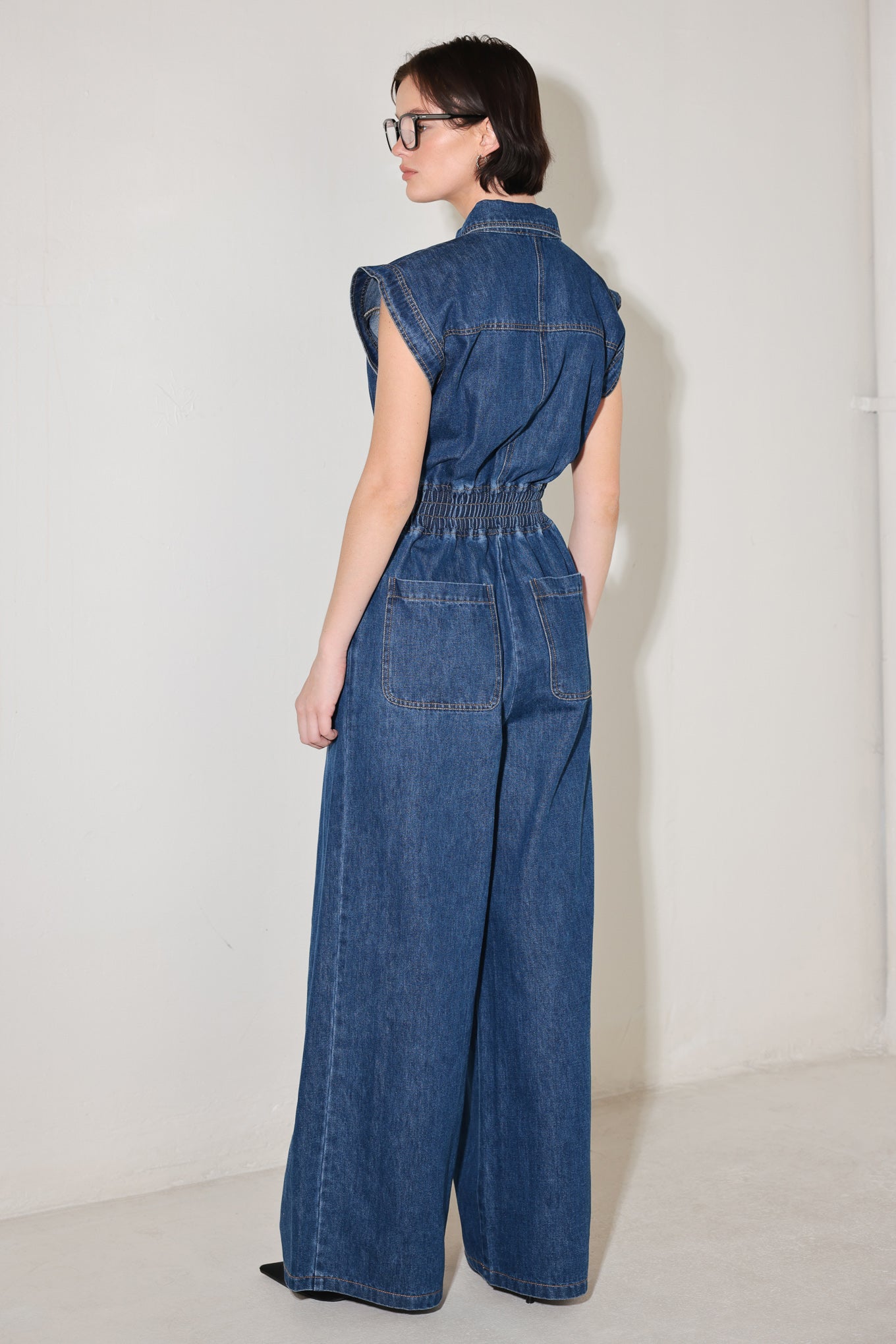 
A washed denim jumpsuit featuring collar, short sleeve, front button down, smocked waistband, side pockets, front pleats and wide leg

Details:

Self : 80% Cotton 20% Polyester

Size &amp; Fit

- Model is 5`8" And Wearing Size Small
- Measurements Taken From Size Small
- Approx. Length: 60"