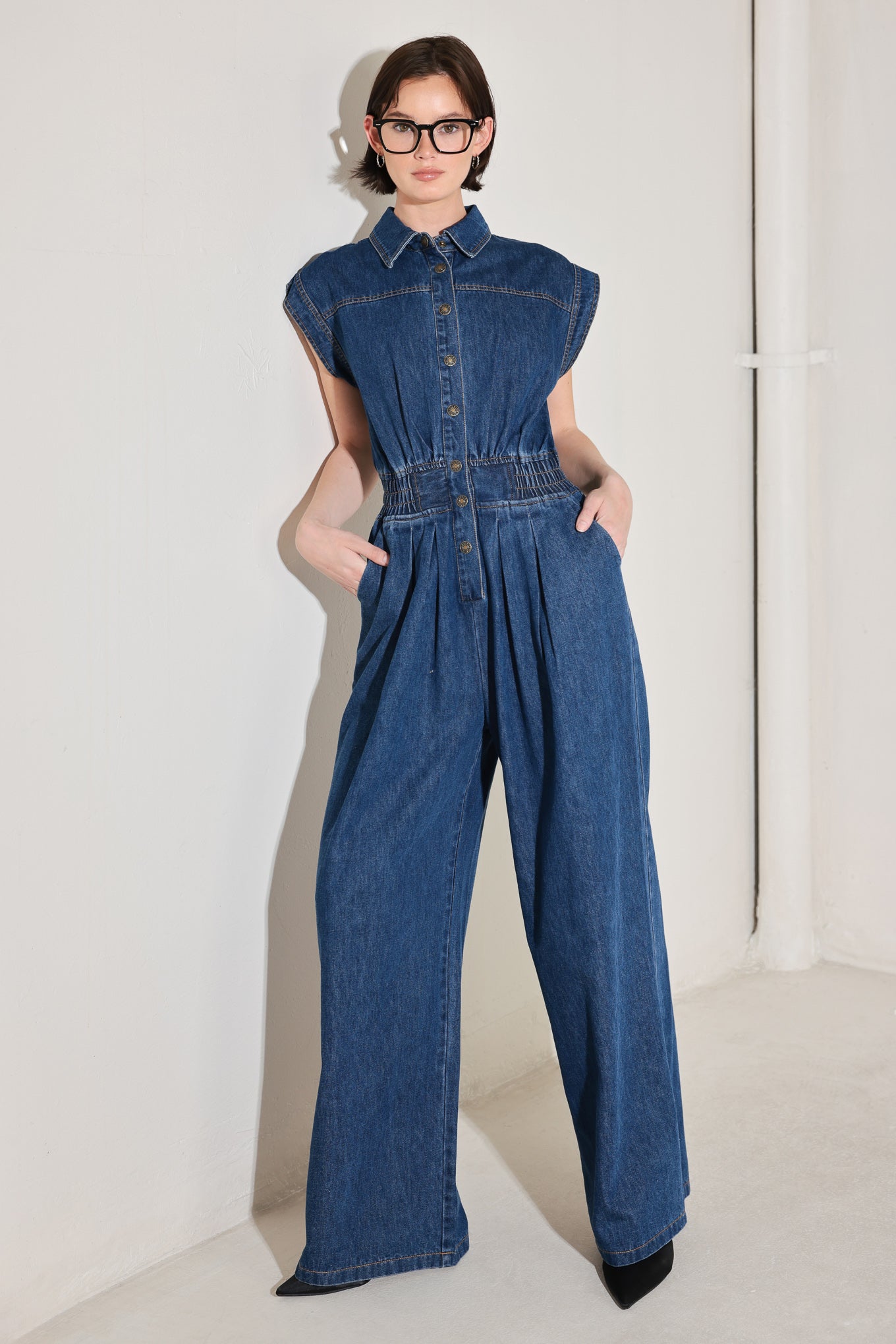 
A washed denim jumpsuit featuring collar, short sleeve, front button down, smocked waistband, side pockets, front pleats and wide leg

Details:

Self : 80% Cotton 20% Polyester

Size &amp; Fit

- Model is 5`8" And Wearing Size Small
- Measurements Taken From Size Small
- Approx. Length: 60"