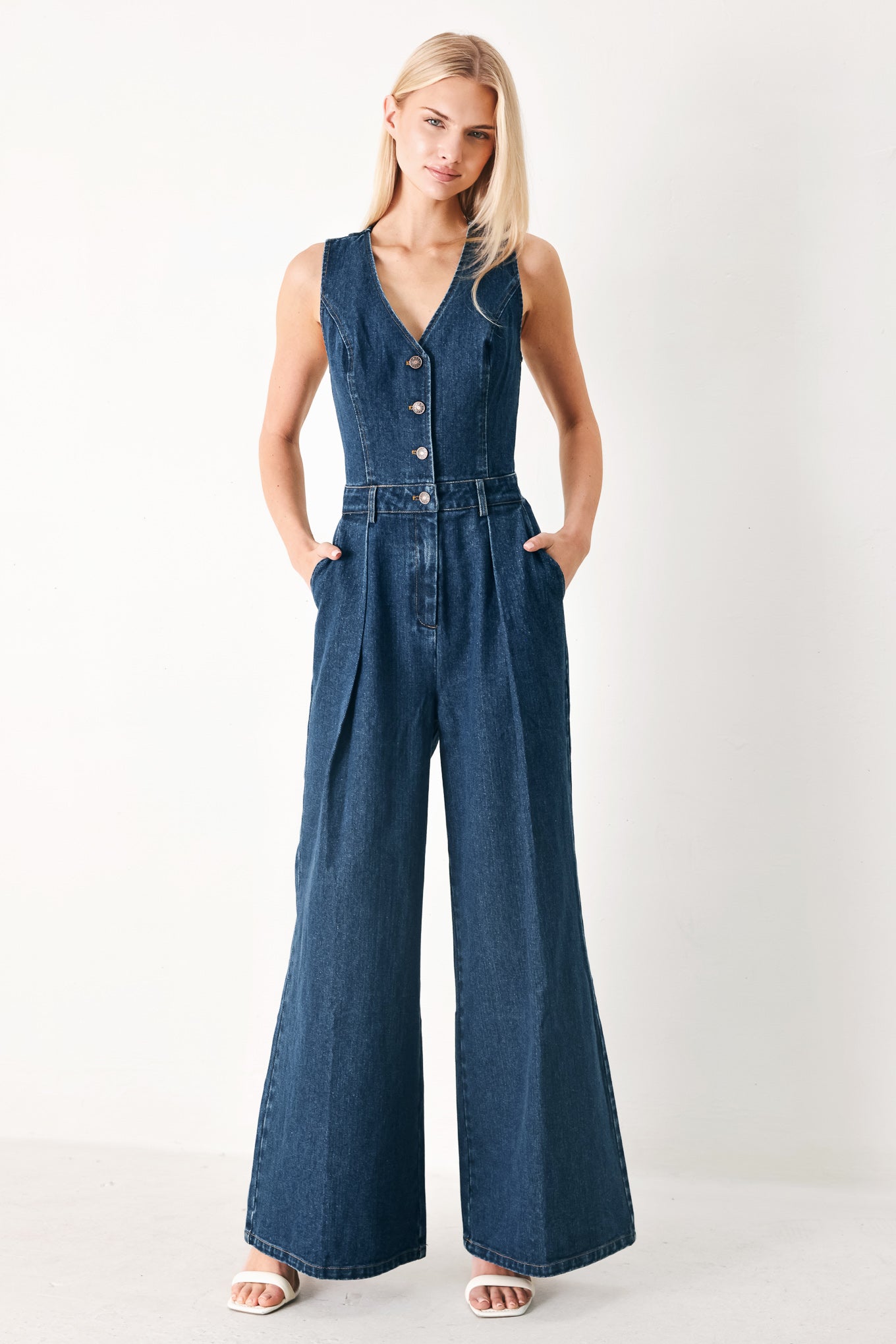 Woman wearing a denim jumpsuit featuring V neckline, sleeveless, front button closure, side pockets, wide leg and surplice back with cut out | FREEMARKET