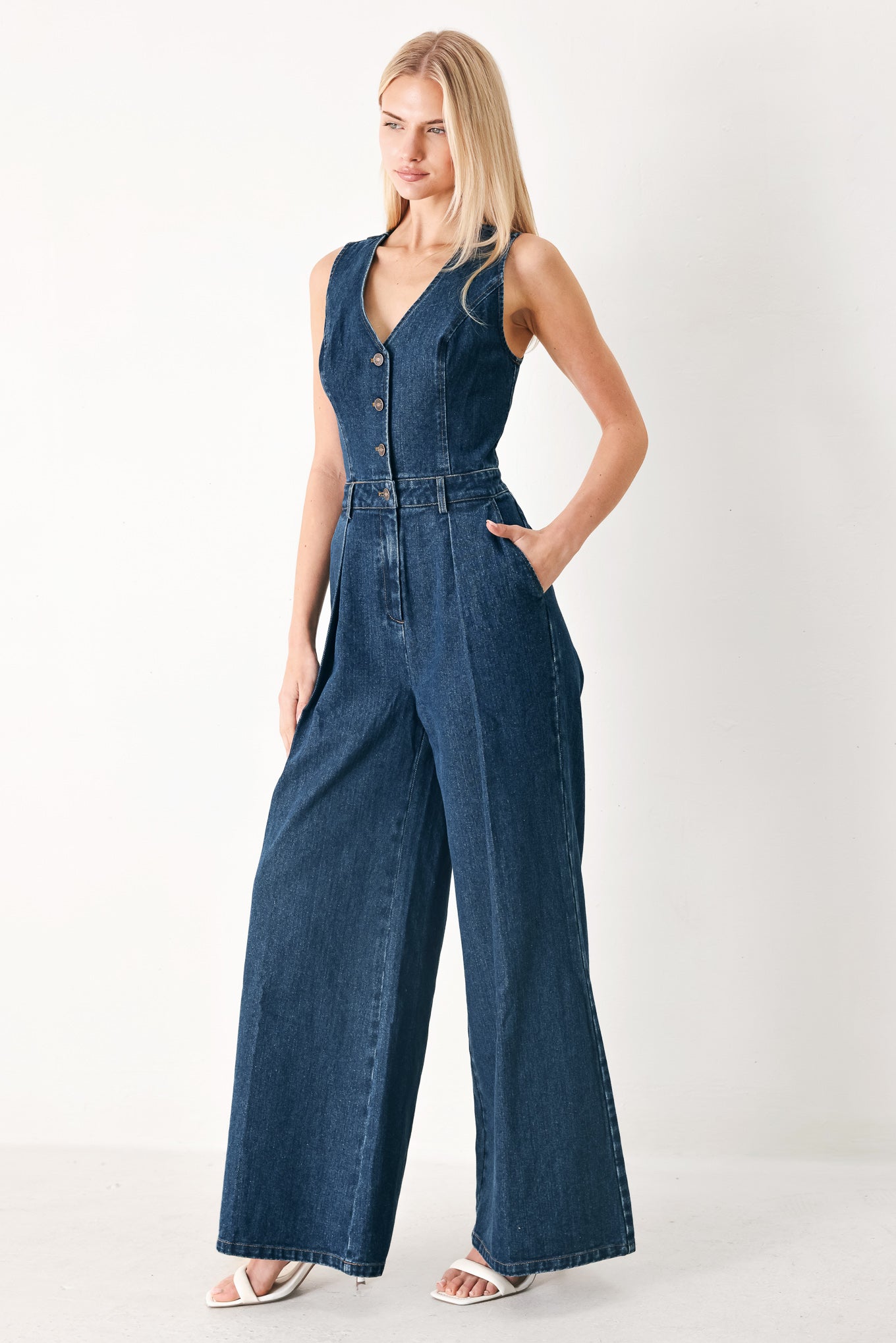Woman wearing a denim jumpsuit featuring V neckline, sleeveless, front button closure, side pockets, wide leg and surplice back with cut out | FREEMARKET