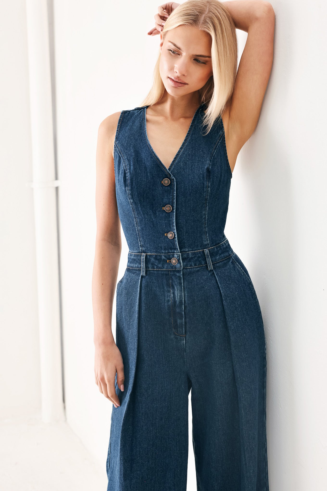 Woman wearing a denim jumpsuit featuring V neckline, sleeveless, front button closure, side pockets, wide leg and surplice back with cut out | FREEMARKET