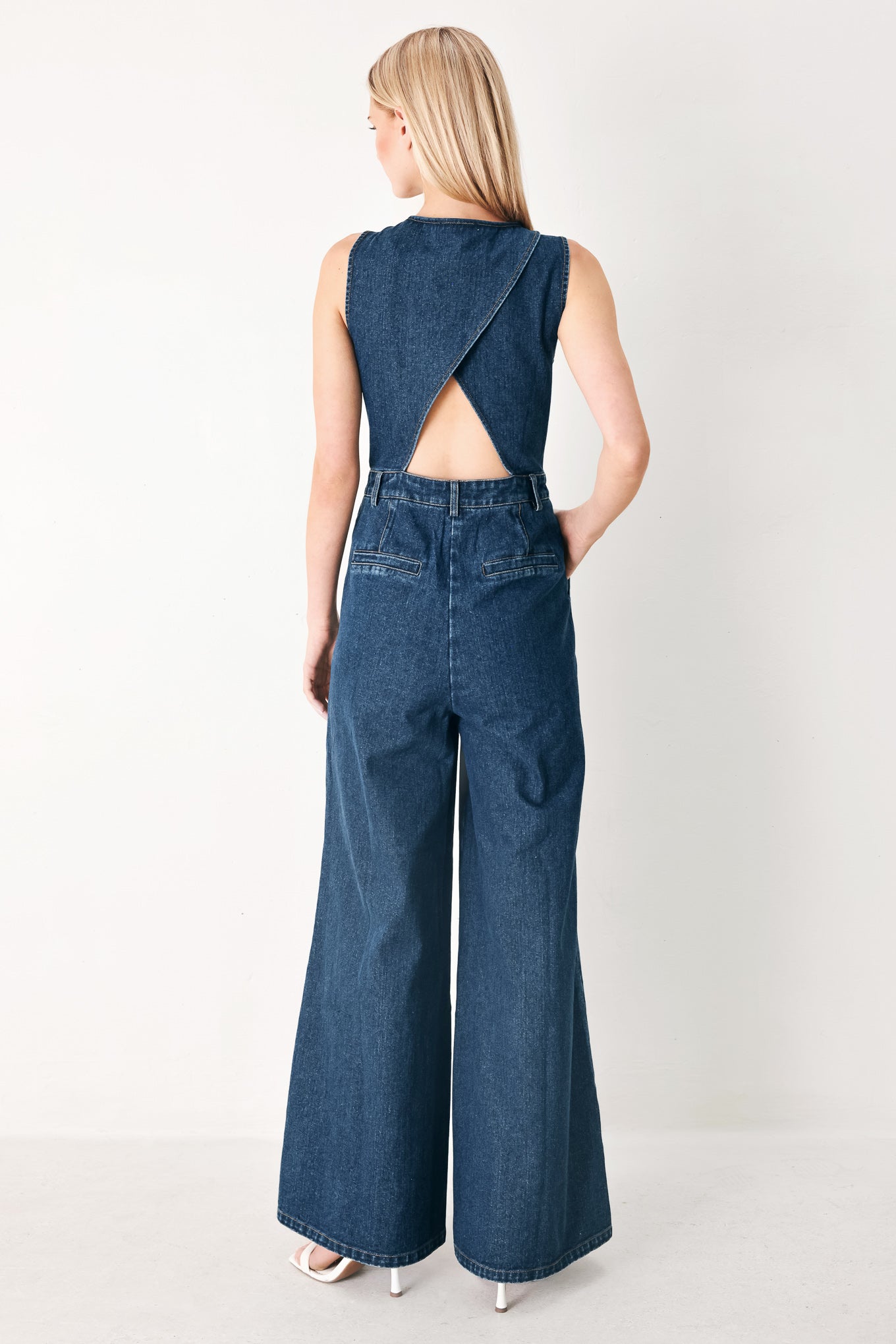 Woman wearing a denim jumpsuit featuring V neckline, sleeveless, front button closure, side pockets, wide leg and surplice back with cut out | FREEMARKET