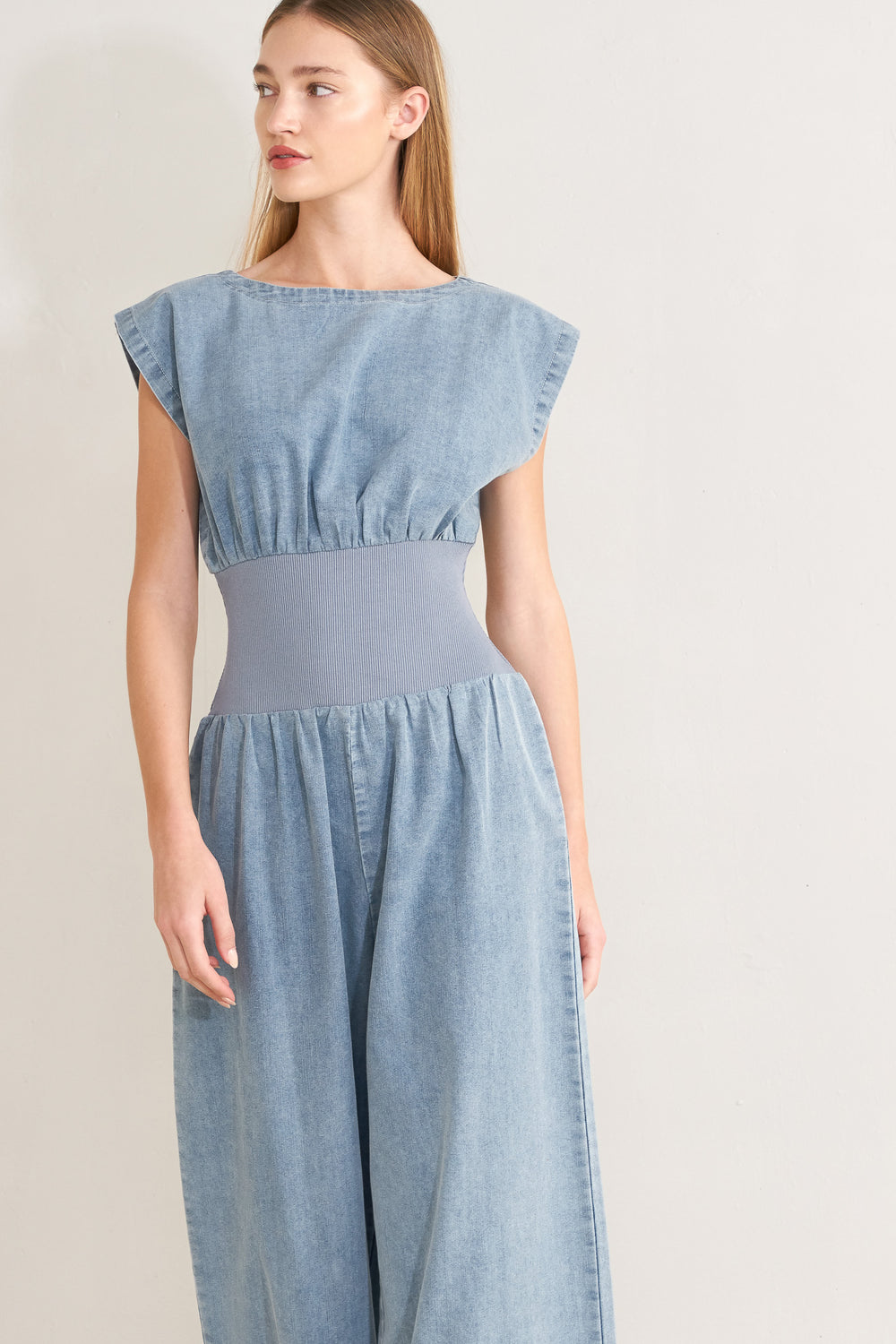 Woman wearing a denim jumpsuit featuring wide neckline, short sleeve, wide knit waistband, wide leg and back slit with button closure | FREEMARKET