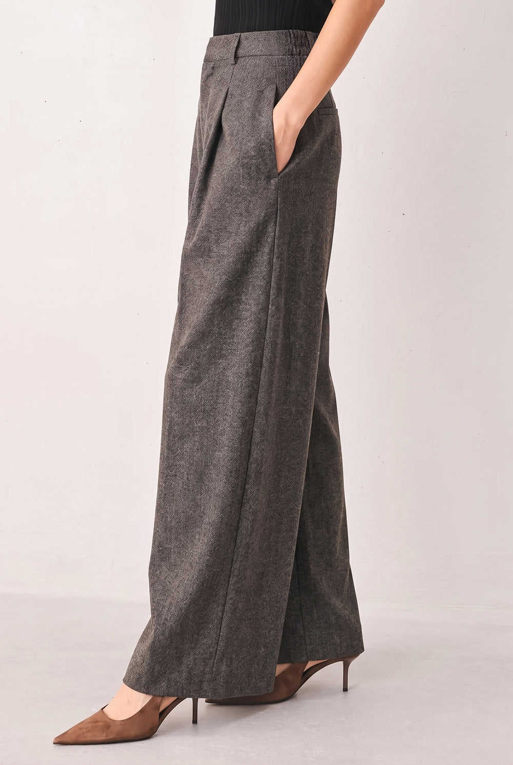 A woman wearing taupe-gray wide-leg pants stands in a styled room holding a laptop, with a clothing rack and fashion magazine pages in the background | FREEMARKET