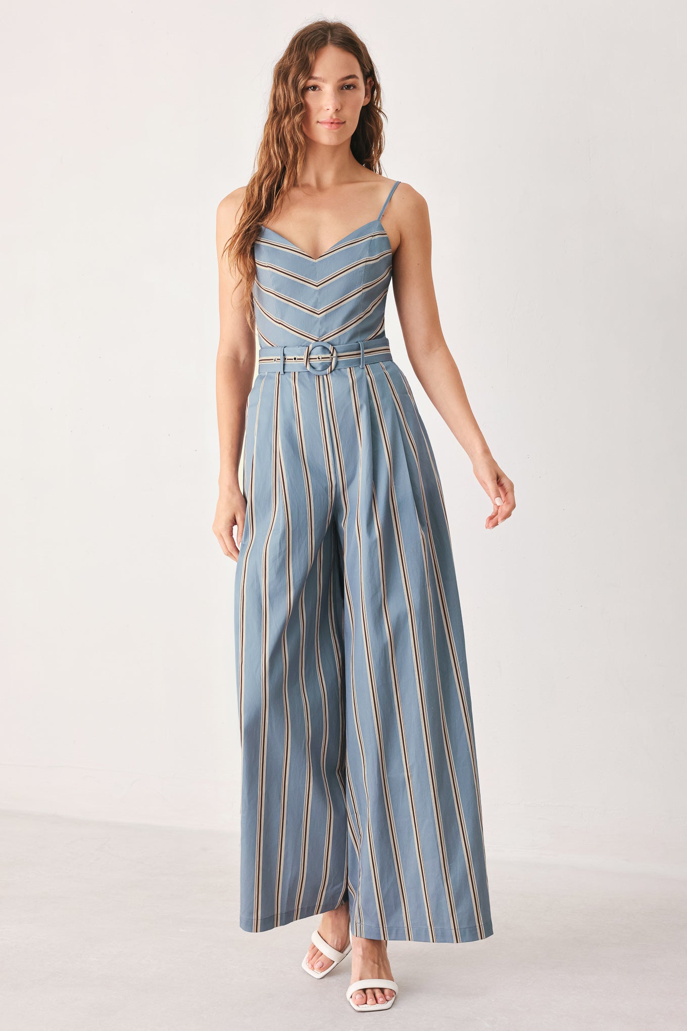 A woman wearing a sleeveless wide-leg jumpsuit with vertical blue, beige, and white stripes, featuring a V-neck bodice, thin shoulder straps, and a matching fabric belt with a silver buckle | FREEMARKET