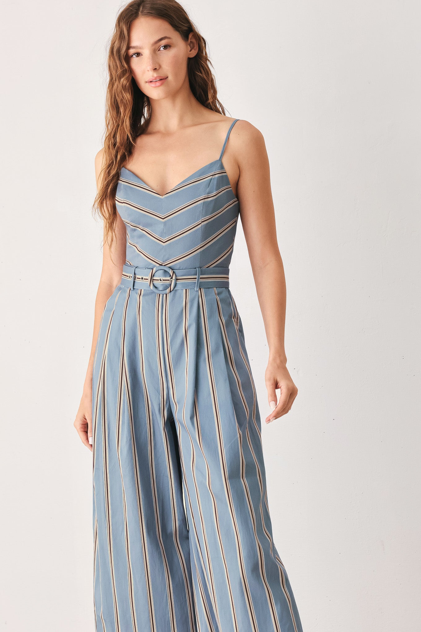 A woman wearing a sleeveless wide-leg jumpsuit with vertical blue, beige, and white stripes, featuring a V-neck bodice, thin shoulder straps, and a matching fabric belt with a silver buckle | FREEMARKET