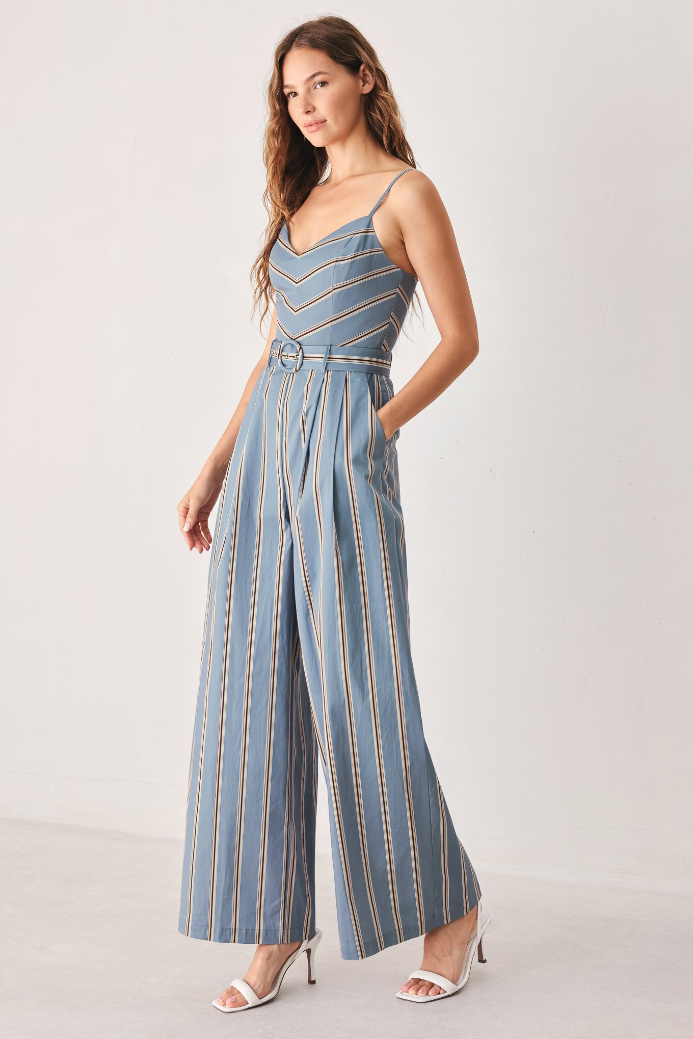 A woman wearing a sleeveless wide-leg jumpsuit with vertical blue, beige, and white stripes, featuring a V-neck bodice, thin shoulder straps, and a matching fabric belt with a silver buckle | FREEMARKET