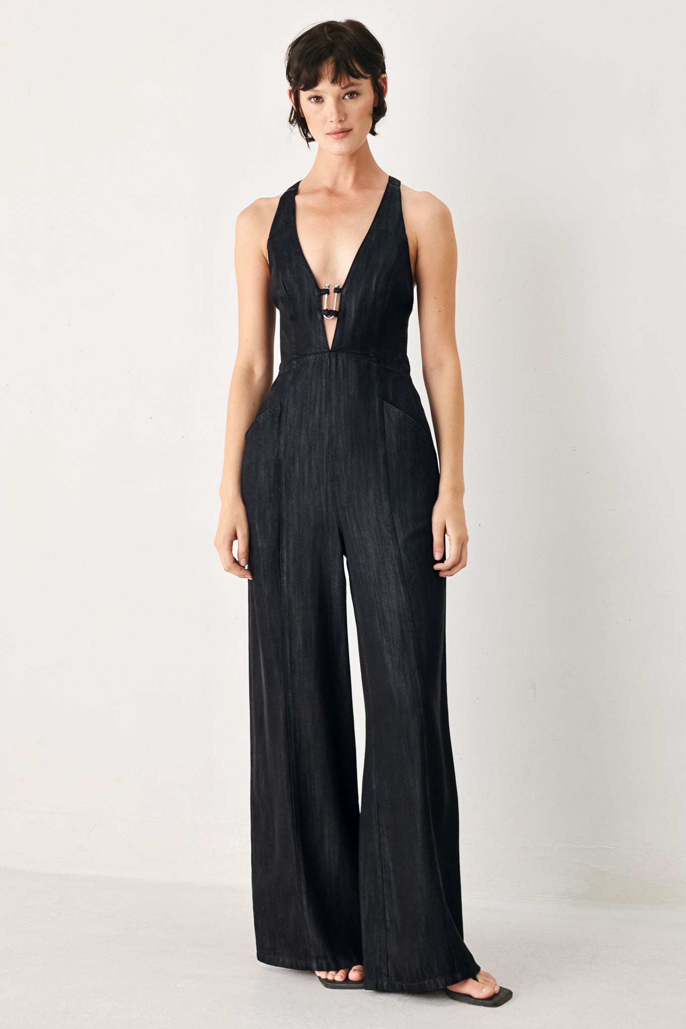 Woman wearing a black denim jumpsuit featuring halter neckline with plunging V and hardware, pockets, wide leg, bare back with cross straps and zipper closure | FREEMARKET