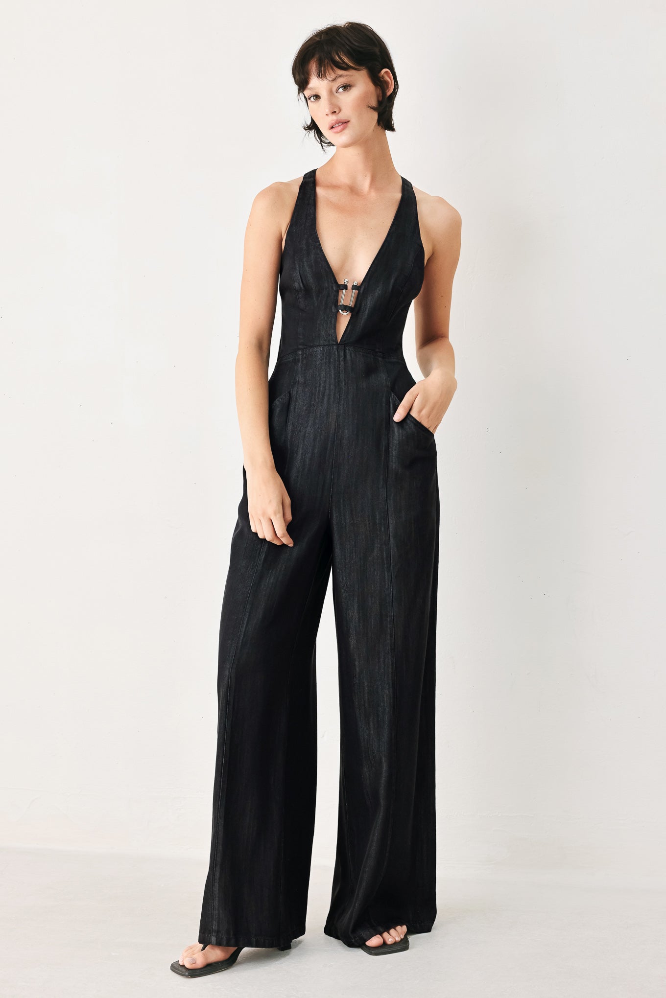 Woman wearing a black denim jumpsuit featuring halter neckline with plunging V and hardware, pockets, wide leg, bare back with cross straps and zipper closure | FREEMARKET
