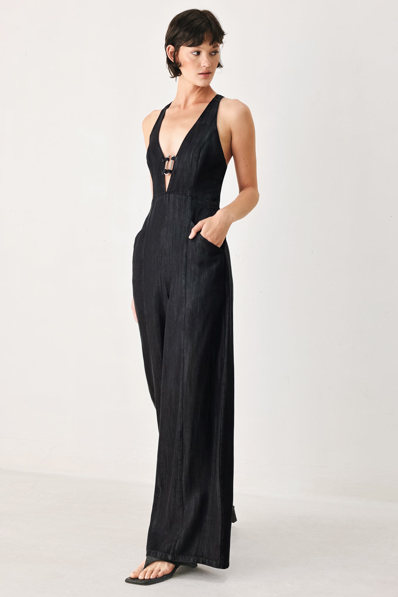 Woman wearing a black denim jumpsuit featuring halter neckline with plunging V and hardware, pockets, wide leg, bare back with cross straps and zipper closure | FREEMARKET