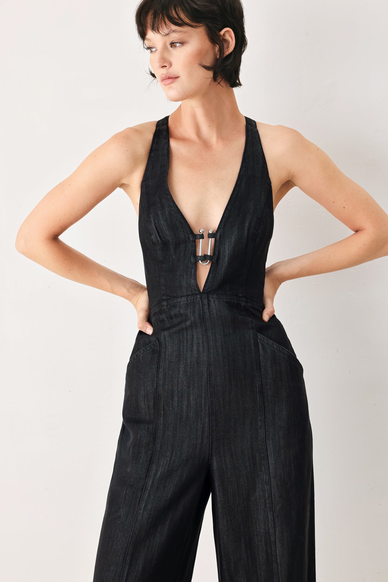 Woman wearing a black denim jumpsuit featuring halter neckline with plunging V and hardware, pockets, wide leg, bare back with cross straps and zipper closure | FREEMARKET