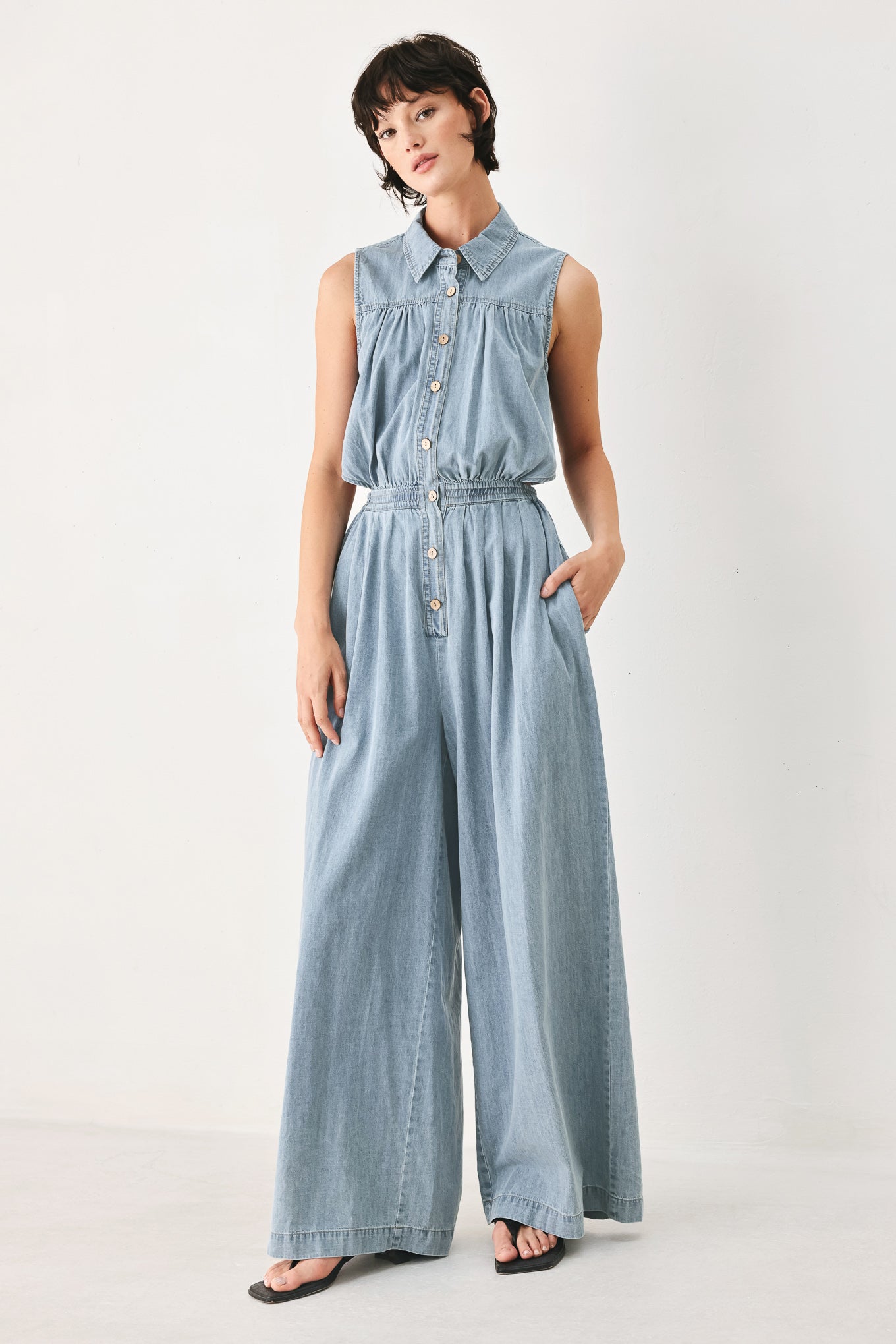 Woman wearing a light blue denim jumpsuit featuring collar, front button down, sleeveless, wide leg, elastic back waistband and popover | FREEMARKET