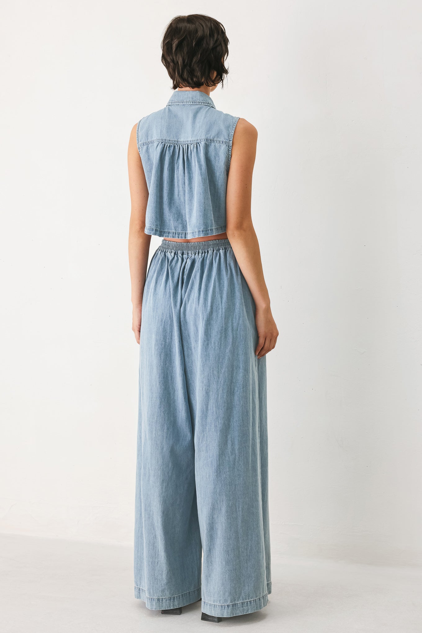 Woman wearing a light blue denim jumpsuit featuring collar, front button down, sleeveless, wide leg, elastic back waistband and popover | FREEMARKET
