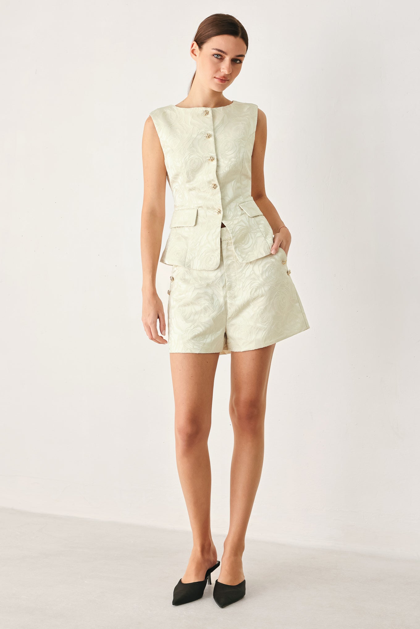 Woman wearing high-waisted cream shorts with a subtle embossed swirl pattern and decorative gold-tone buttons down the side pocket | FREEMARKET