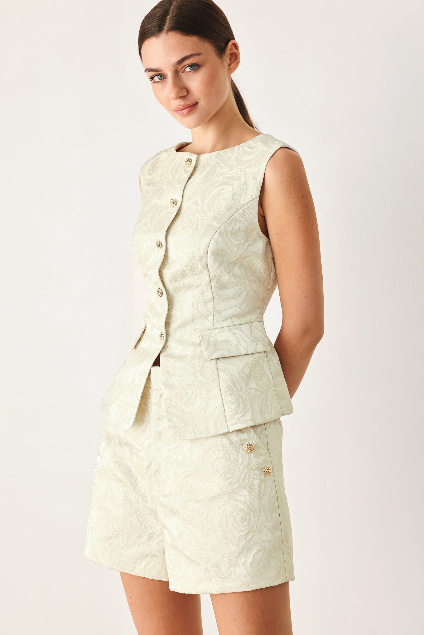 Woman wearing high-waisted cream shorts with a subtle embossed swirl pattern and decorative gold-tone buttons down the side pocket | FREEMARKET