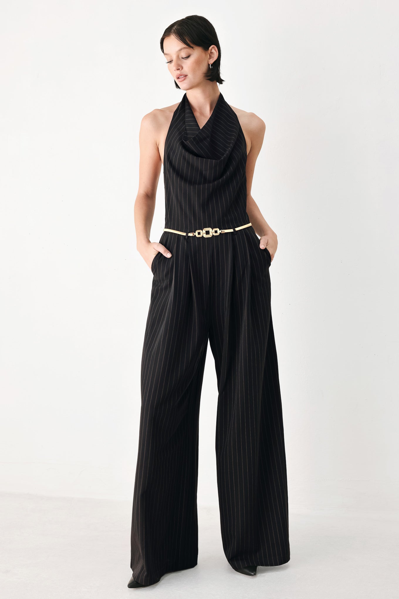 A model wearing a black pinstriped halter-neck wide-leg jumpsuit with a gold chain belt | FREEMARKET