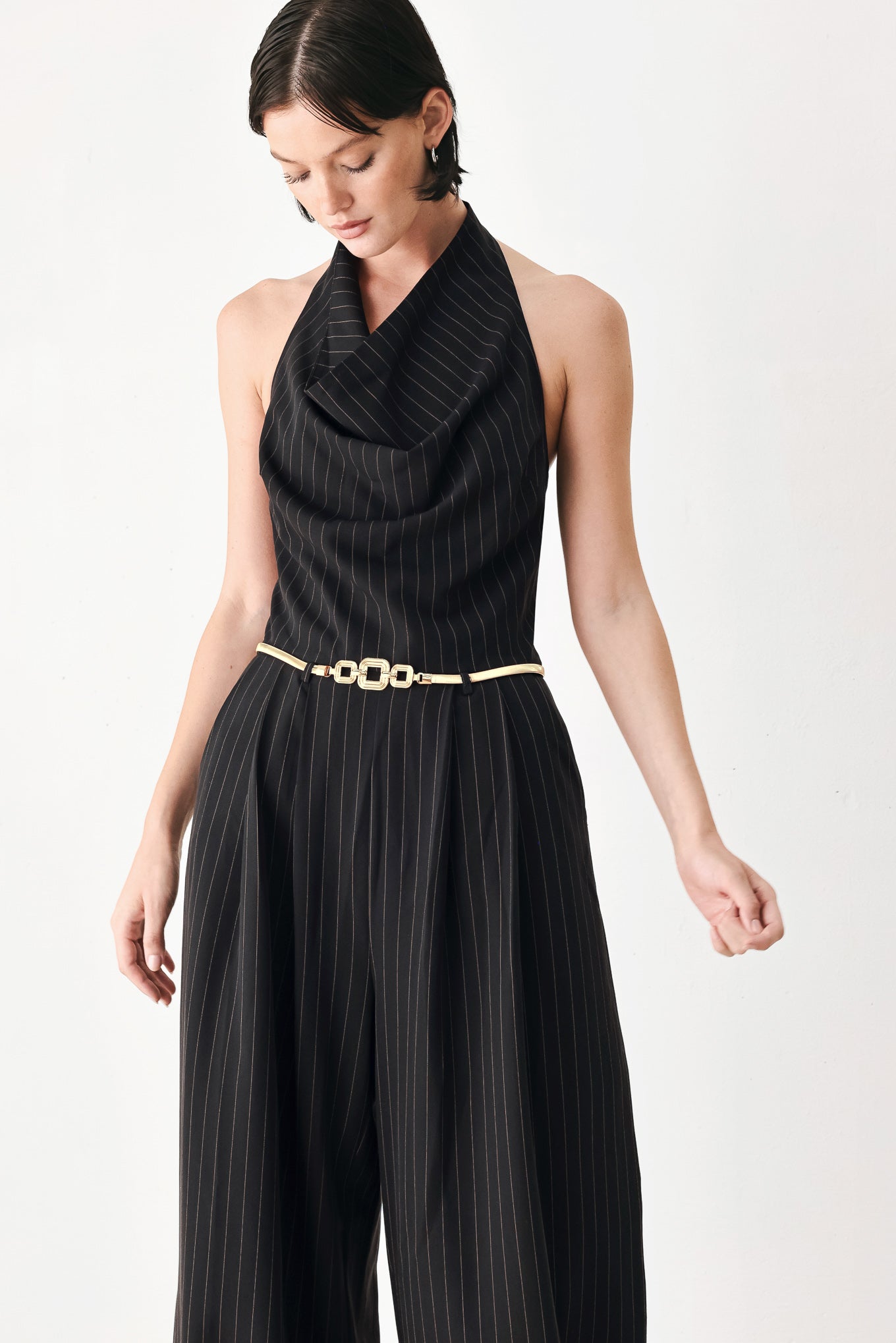 A model wearing a black pinstriped halter-neck wide-leg jumpsuit with a gold chain belt | FREEMARKET