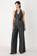 A model wearing a sleeveless halter-neck jumpsuit with black and white pinstripes, a matching black belt, and wide-leg trousers, standing against a plain white studio background.