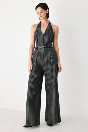 A model wearing a sleeveless halter-neck jumpsuit with black and white pinstripes, a matching black belt, and wide-leg trousers, standing against a plain white studio background.