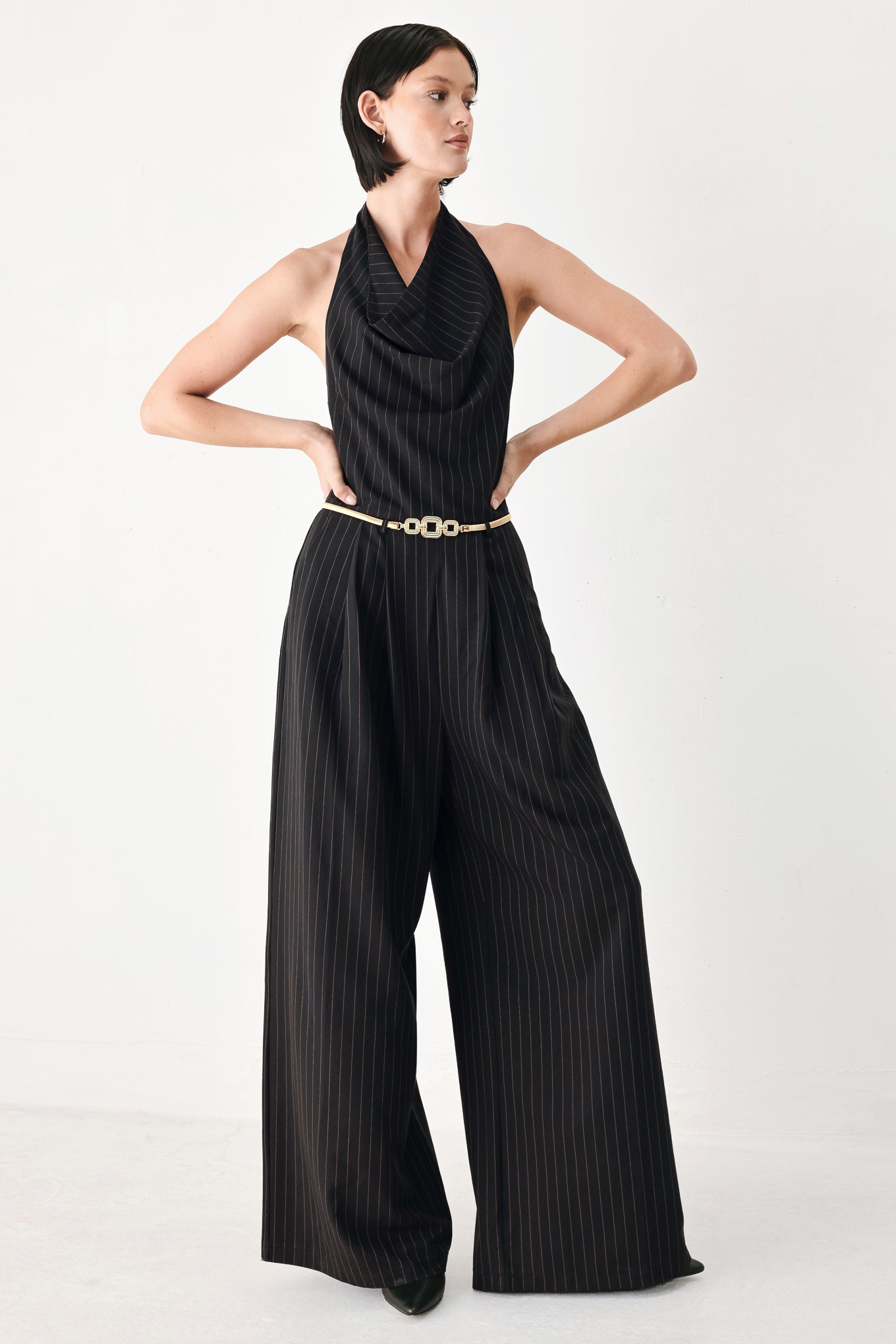 A model wearing a black pinstriped halter-neck wide-leg jumpsuit with a gold chain belt | FREEMARKET