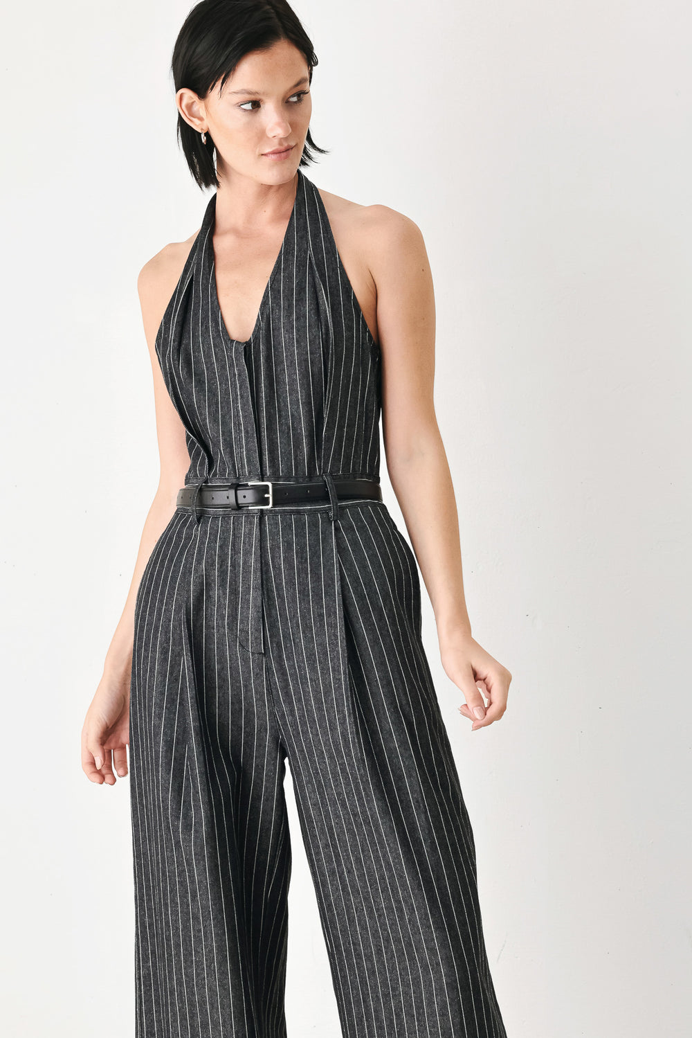 A model wearing a sleeveless halter-neck jumpsuit with black and white pinstripes, a matching black belt, and wide-leg trousers, standing against a plain white studio background.