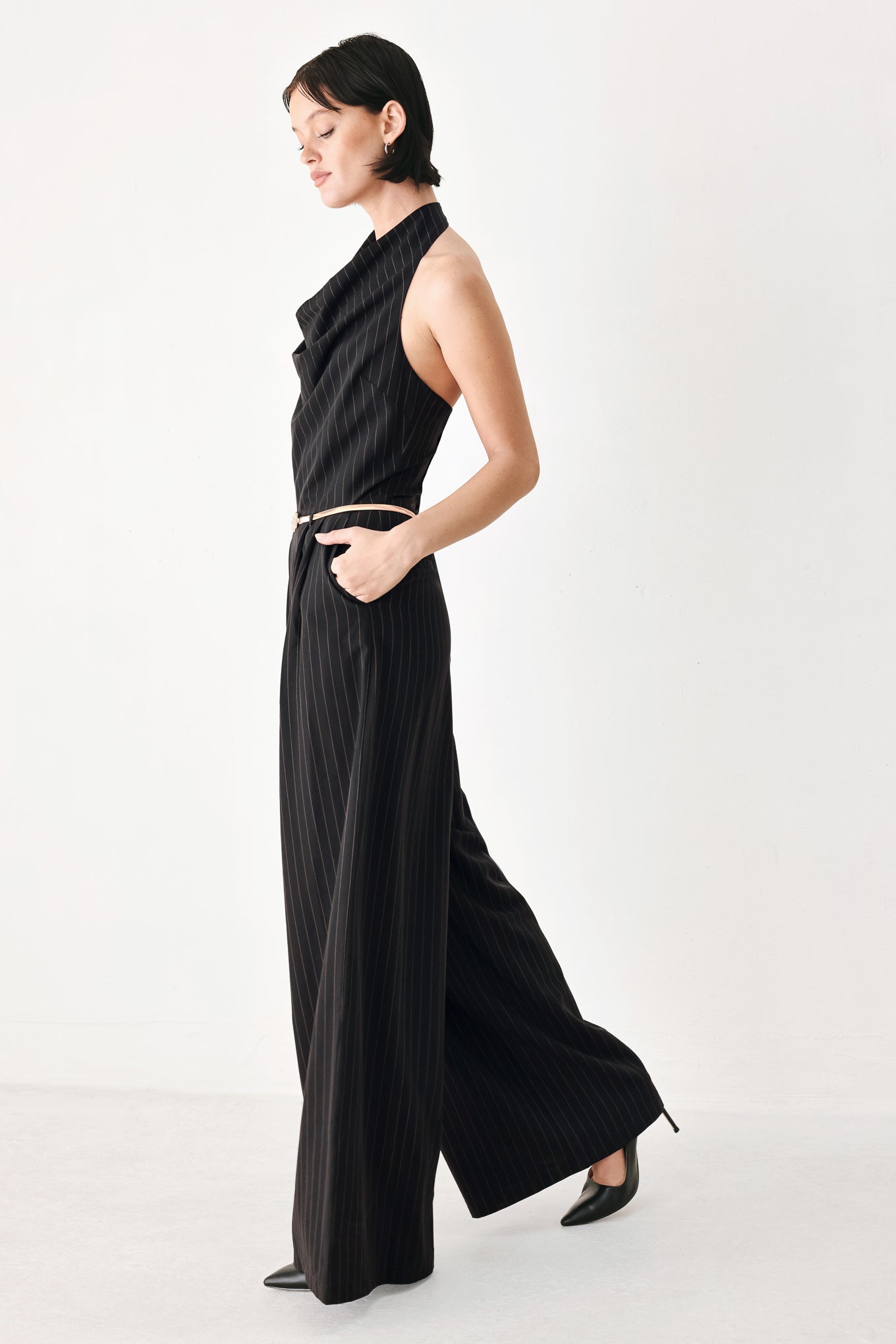 A model wearing a black pinstriped halter-neck wide-leg jumpsuit with a gold chain belt | FREEMARKET