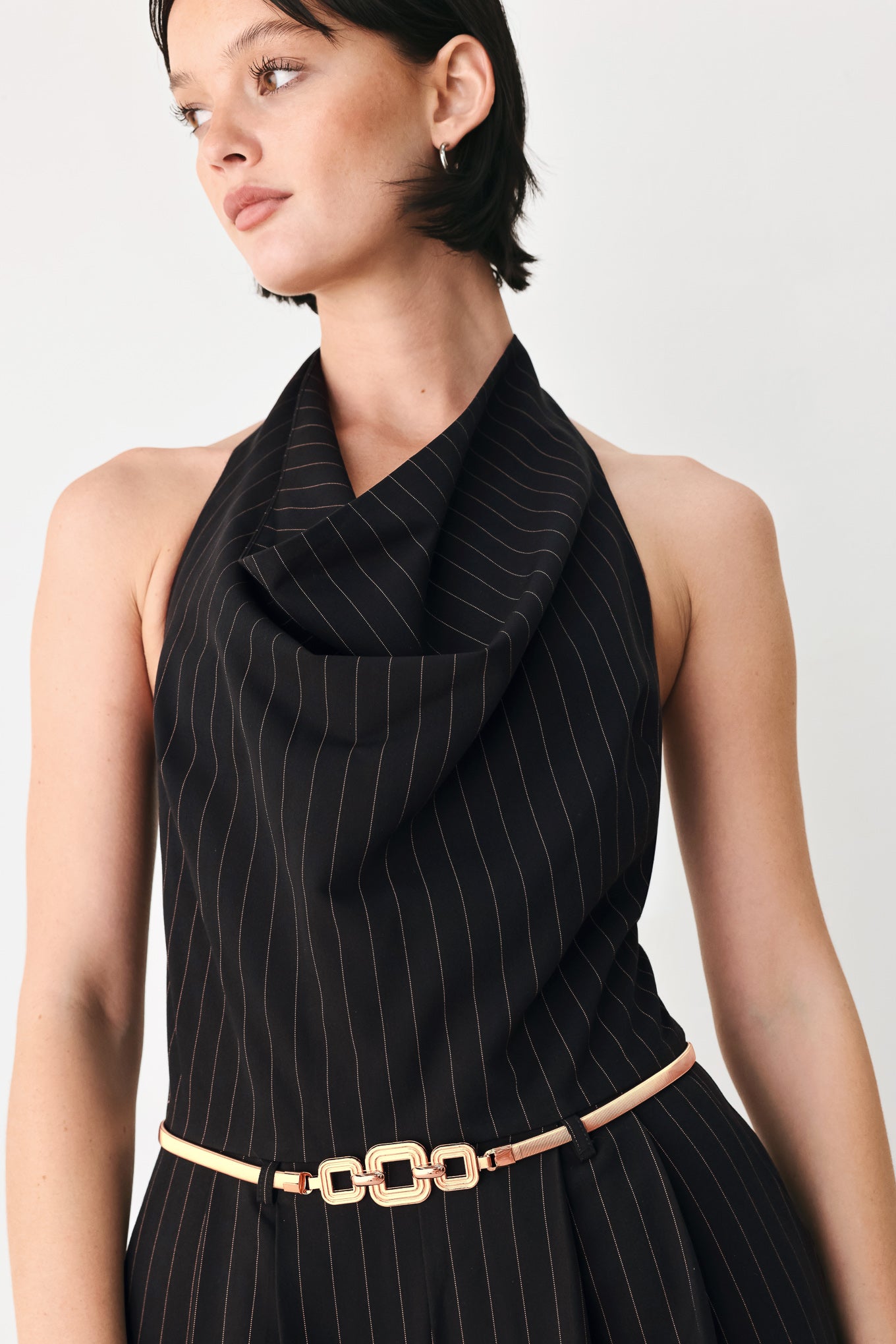 A model wearing a black pinstriped halter-neck wide-leg jumpsuit with a gold chain belt | FREEMARKET