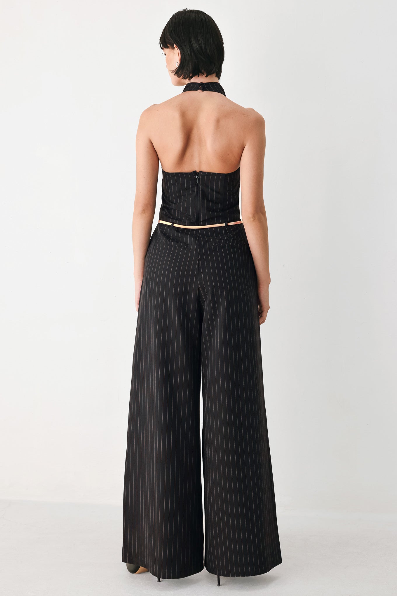 A model wearing a black pinstriped halter-neck wide-leg jumpsuit with a gold chain belt | FREEMARKET