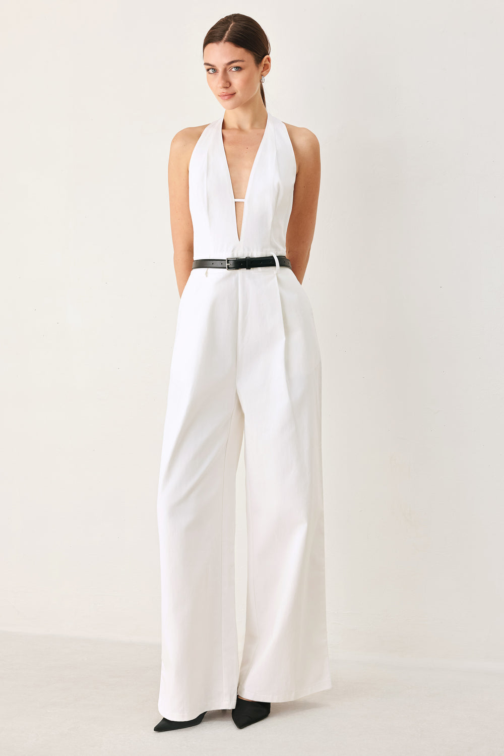 Woman wearing a white woven jumpsuit featuring halter neckline with plunging V front, contrasting belt, single pleat pant with side pocket and bare back with zipper closure | FREEMARKET