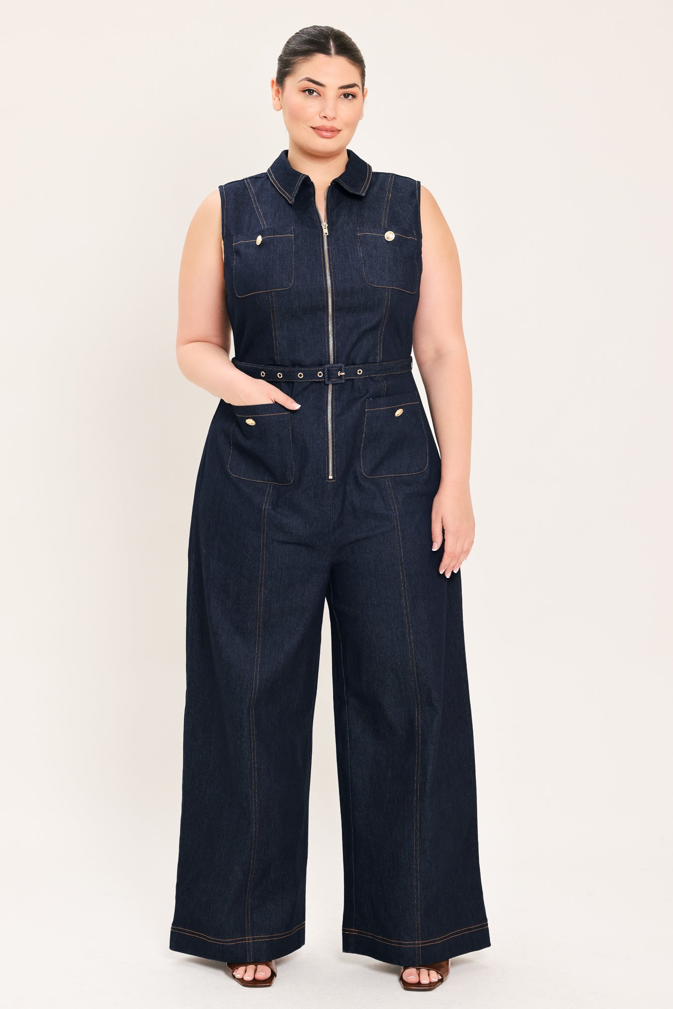 Woman wearing a dark blue denim sleeveless, wide-leg jumpsuit featuring collar, front zipper closure, and pockets | Plus Curve  FREEMARKET